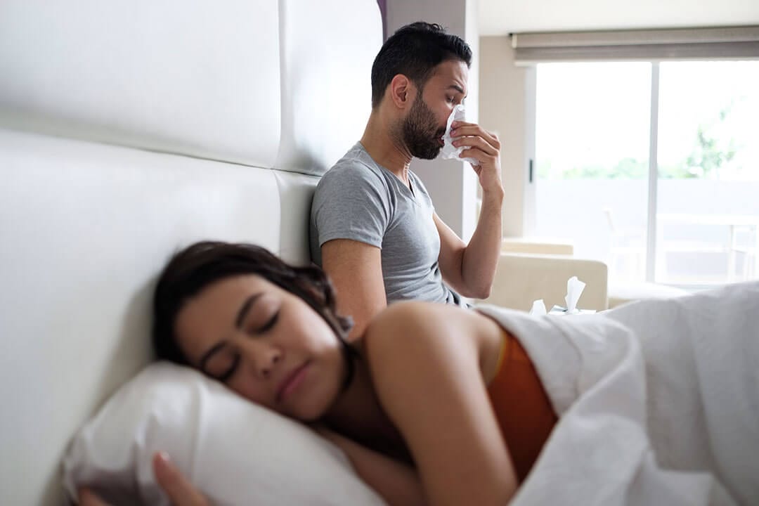 Young Man with Allergies Blows Nose in Bed While Female Partner Sleeps