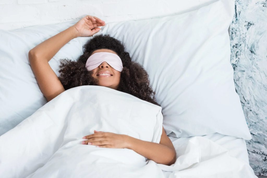 Young Black woman laying happily in bed wearing an eye mask.
